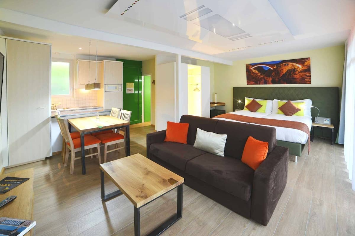 Een moderne hotelkamer met een houten eettafel, een bank met oranje kussens, een bed met veel kussens, en een groot schilderij aan de muur.