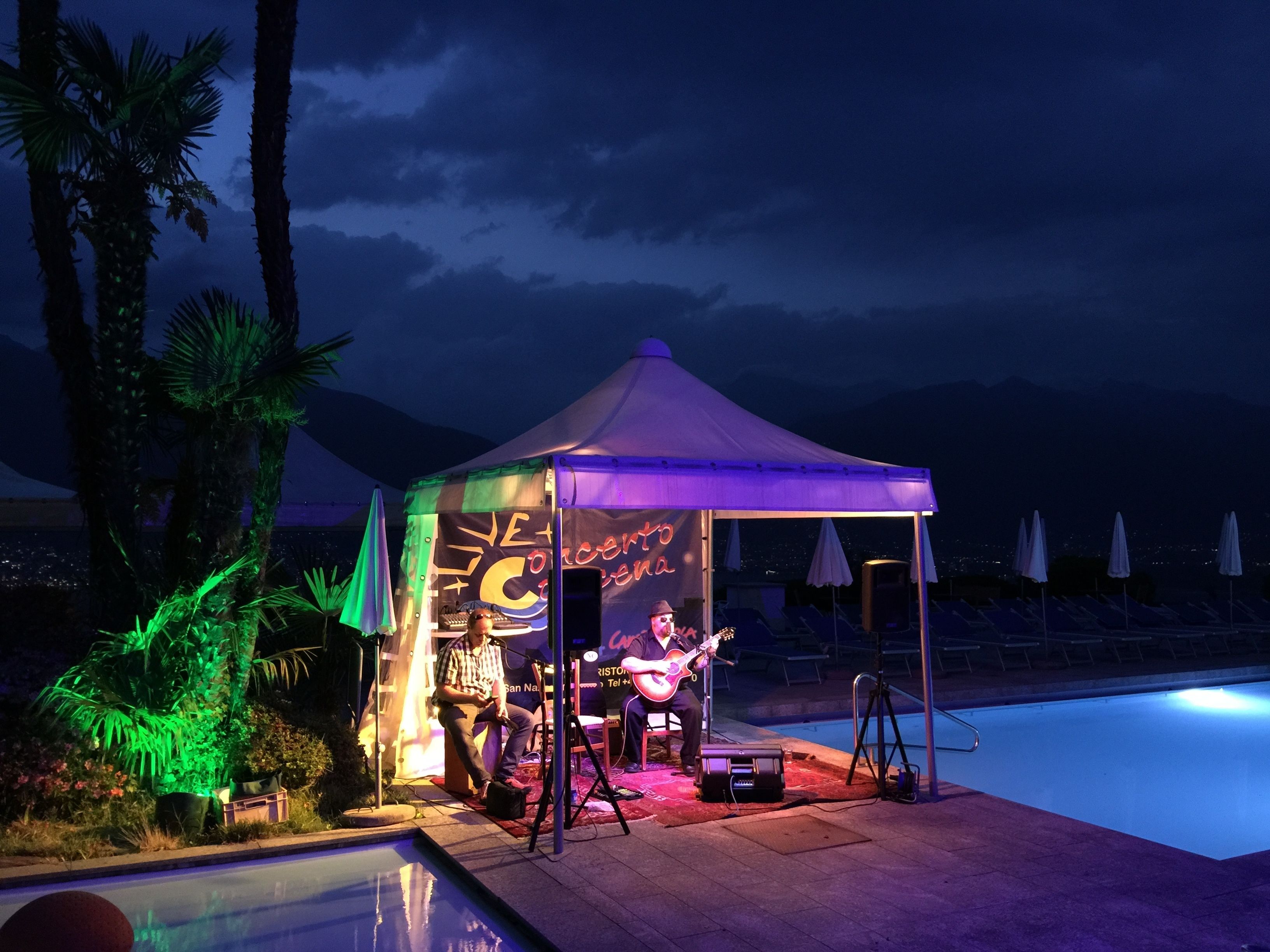 Una banda ofrece un espectáculo de música en vivo al aire libre por la noche, bajo una carpa, cerca de una piscina, con palmeras y montañas al fondo.
