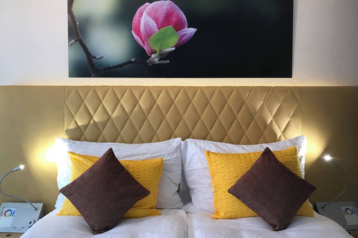 Hotelkamer met een gestoffeerd hoofdeinde, witte en gele kussens, en een lijst met een roze bloem aan de muur.