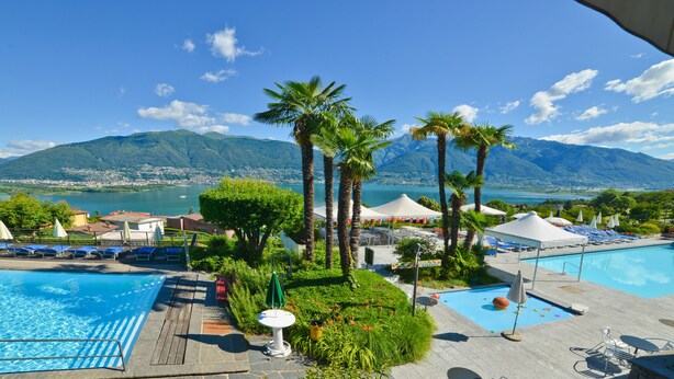 A pool area with two separate pools, surrounded by palm trees and lounge chairs, with a mountain and lake view in the background. - Hotel La Campagnola (Gambarogno)
