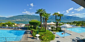 A pool area with two separate pools, surrounded by palm trees and lounge chairs, with a mountain and lake view in the background. - Hotel La Campagnola (Gambarogno)