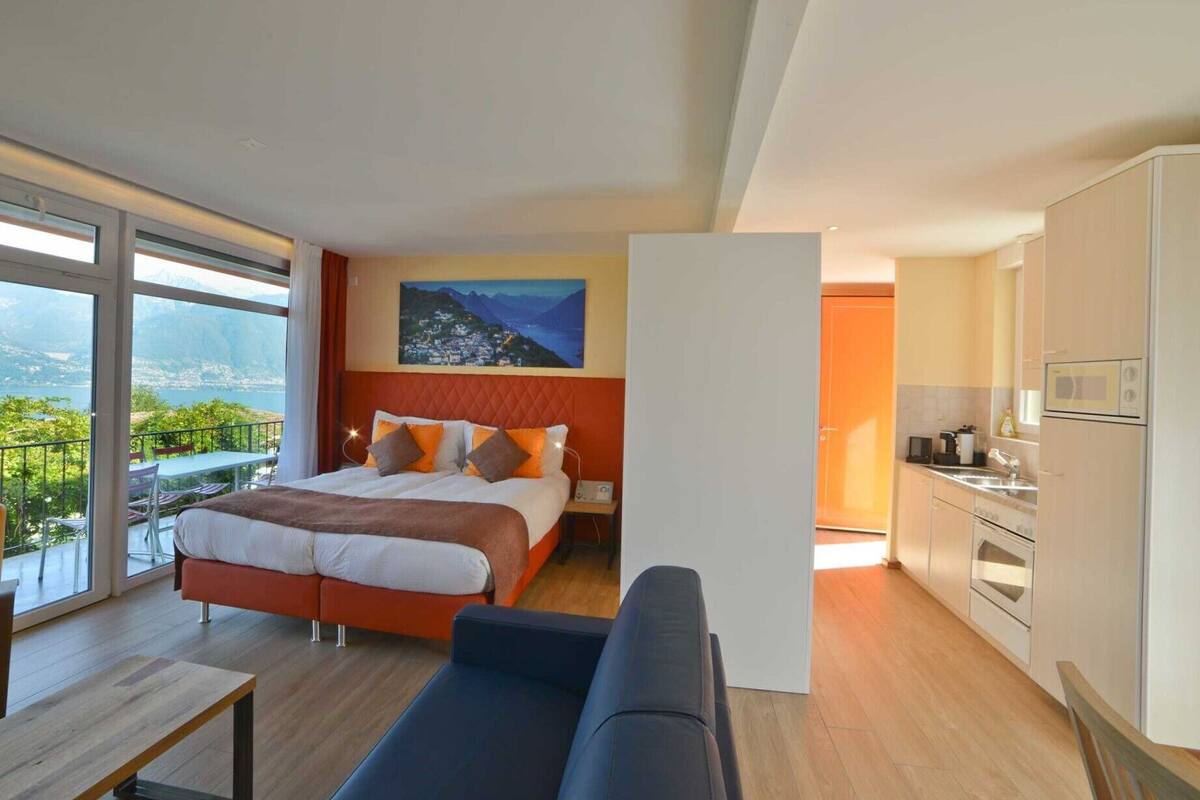 Een moderne hotelkamer met een groot bed, uitzicht op de bergen, een balkon met tuinmeubilair en een kitchenette.
