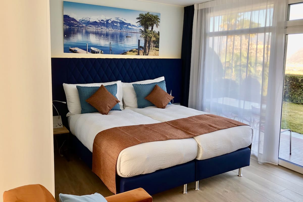 Een moderne slaapkamer met een groot bed, een schilderij van een meer en bergen, en uitzicht op het landschap door dunne gordijnen.