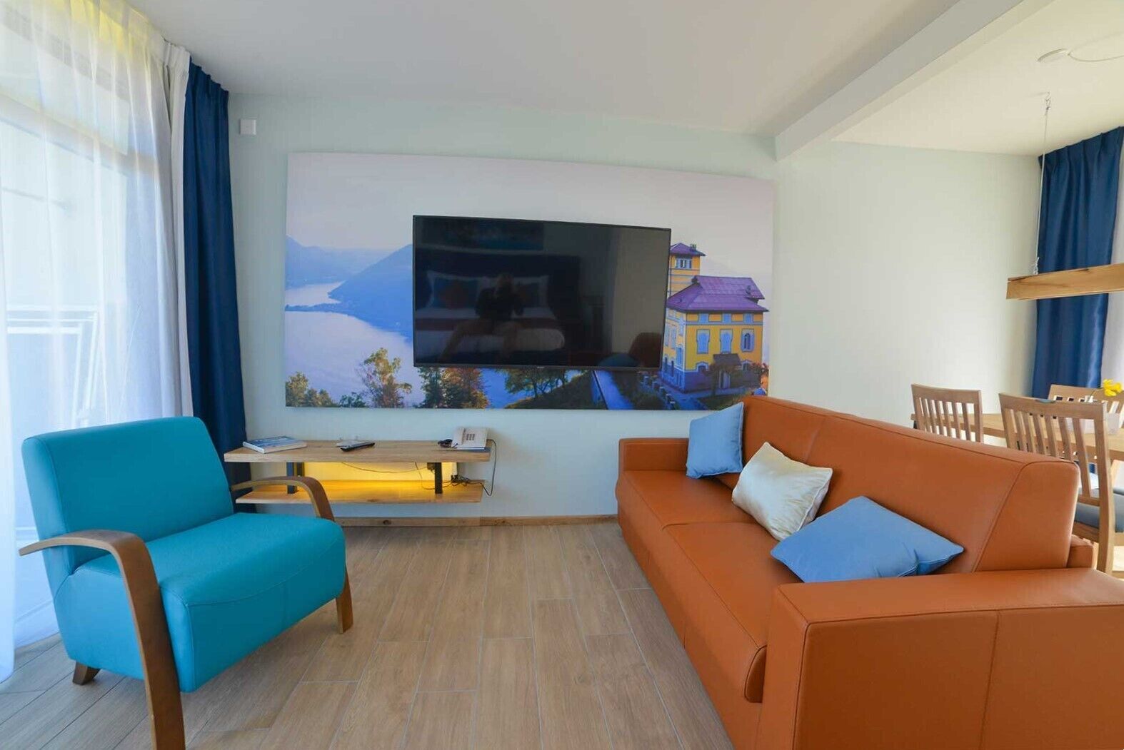 A living room with a large flat-screen TV, a blue armchair, an orange sofa with blue cushions, a wooden coffee table, and a dining area with wooden chairs and a small table.