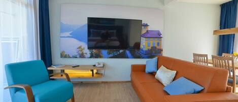 A living room with a large flat-screen TV, a blue armchair, an orange sofa with blue cushions, a wooden coffee table, and a dining area with wooden chairs and a small table.