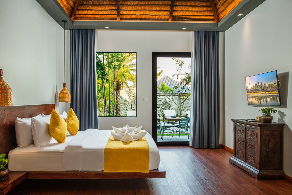 A bedroom with a bed, a wooden headboard, a bedside table with a plant, a large window with curtains, and a view of an outdoor area.