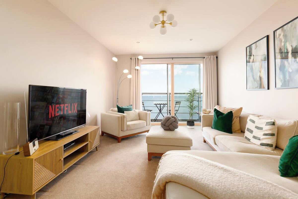 A modern living room with a flat-screen TV, comfortable seating, a balcony with a table and chairs, and large windows offering a view.