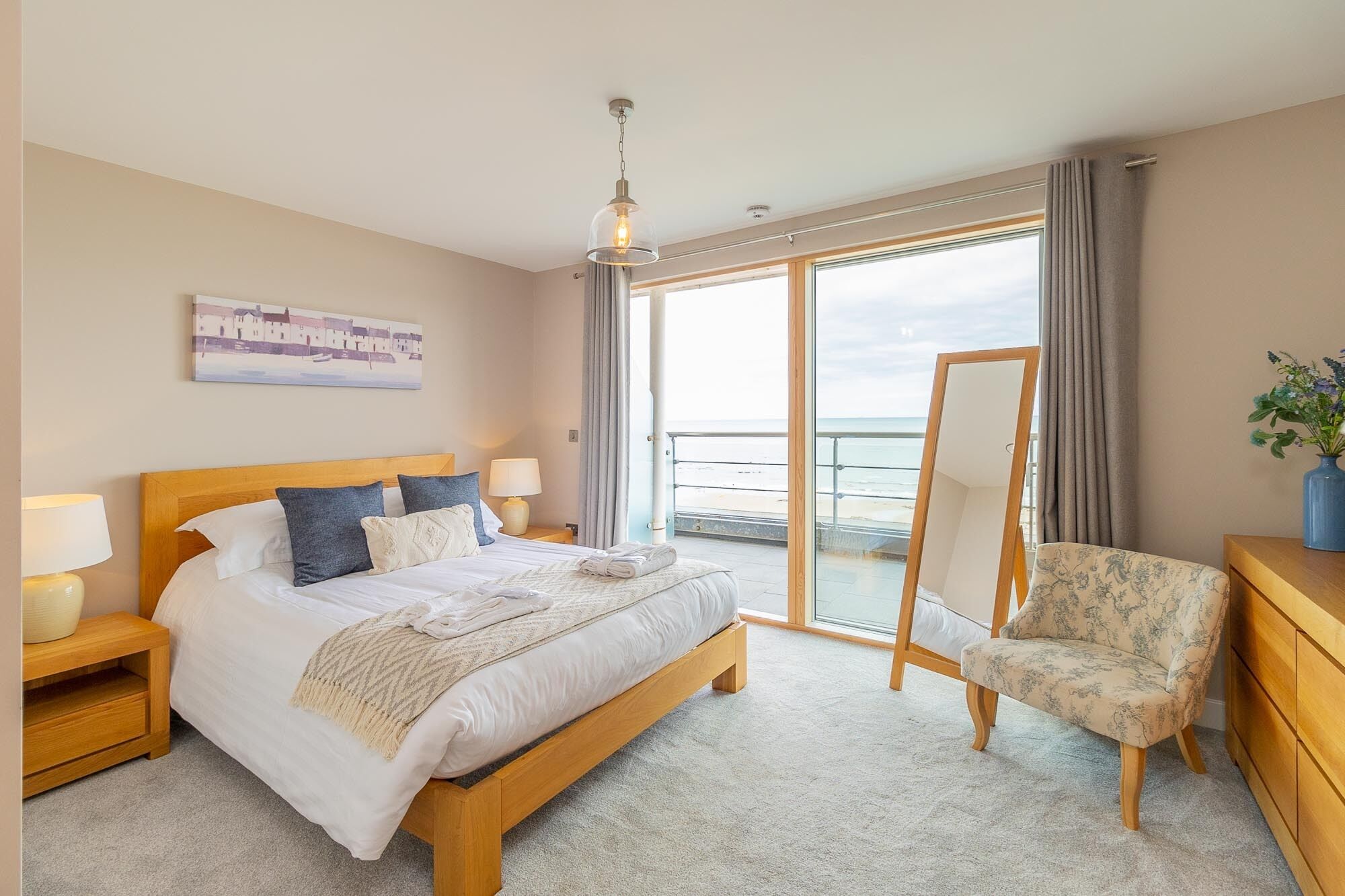 A hotel room with a bed, bedside tables, a chair, a dresser, a painting, and a view of the sea.