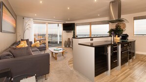 A modern living room with a grey sofa, a coffee table, and a kitchen area with a stainless steel range hood. - The Sands - Sea Front Apartments (Scarborough)