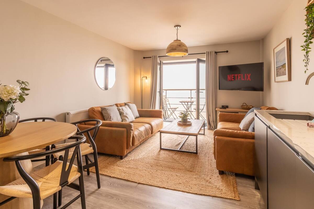 A modern living room with a sofa, coffee table, and dining area. A TV is mounted on the wall displaying 'NETFLIX'.