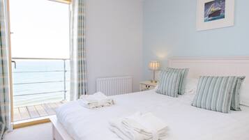 Habitación de hotel con cama, almohadas, mesita de noche, lámpara y vista al mar a través de una puerta abierta.