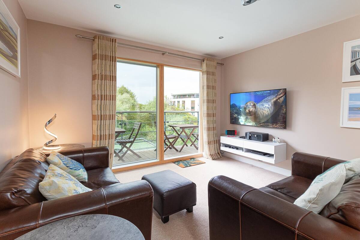 A living room with a brown leather sofa, a television on the wall, and a sliding glass door leading to an outdoor area.