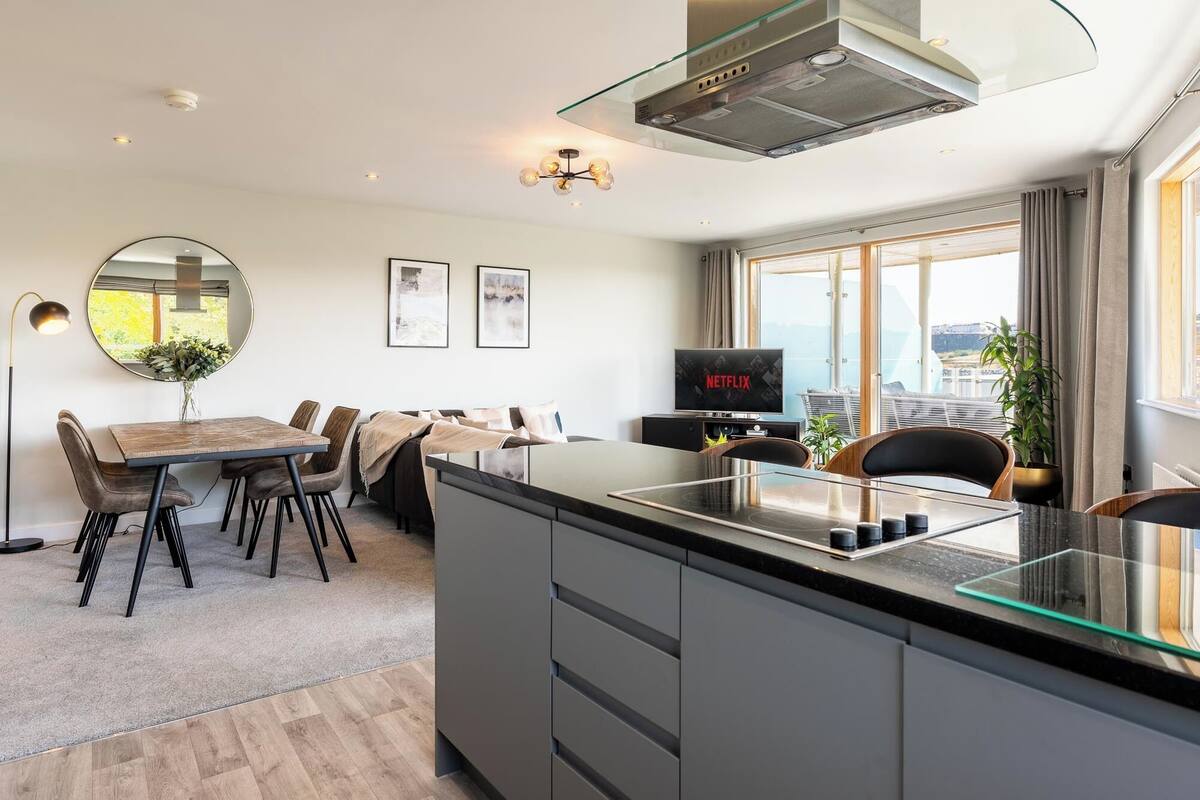 A modern kitchen with a glass island, a dining area with a round mirror, and a TV displaying 'NETFLIX'.