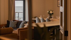 A modern living room with a sofa, a dining table with chairs, and a view of the ocean through a large window. - The Sands - Sea Front Apartments (Scarborough)