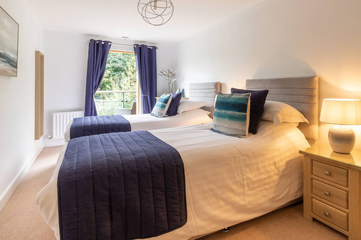 A hotel room with two beds, a window with curtains, a bedside table with a lamp, and a radiator.
