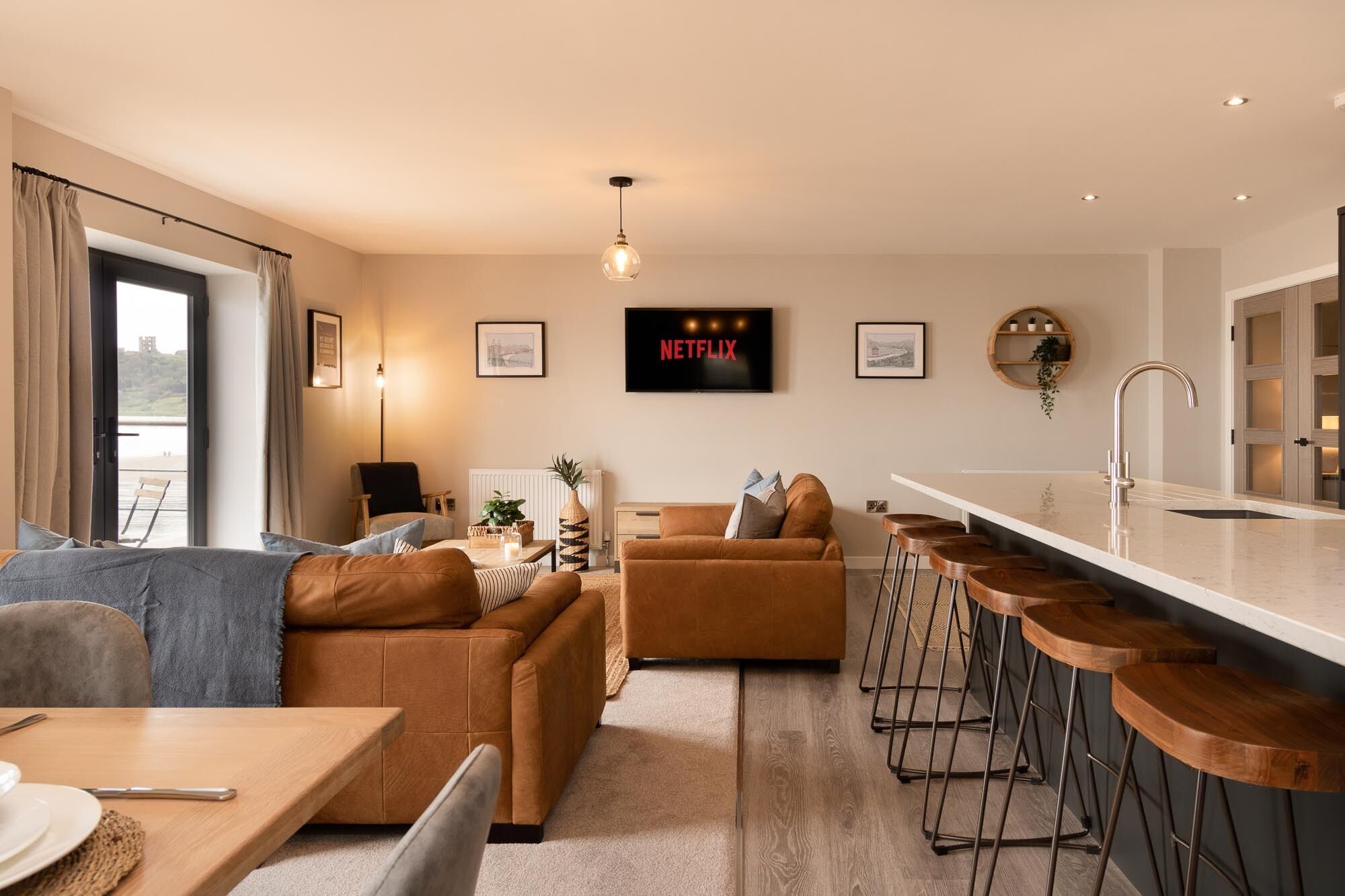 Un salon moderne avec un canapé, une table basse et un téléviseur affichant le logo « NETFLIX ». On y trouve également un coin cuisine avec un bar et des tabourets.