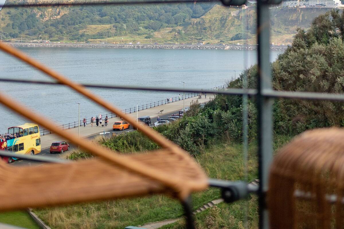 A view from a window overlooking a body of water, a hill with a structure, and a road with vehicles and people.