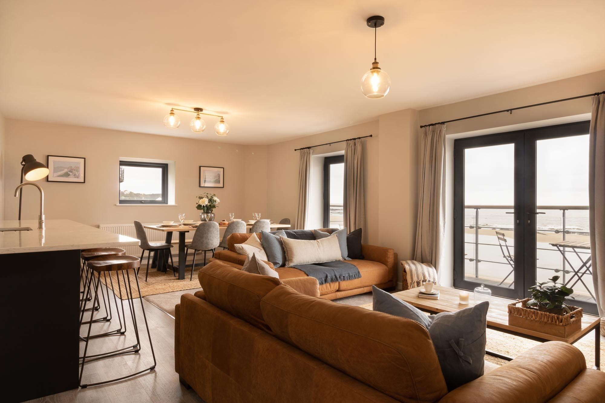 Een moderne woonkamer met een bank, een eethoek en grote ramen met uitzicht op het strand.