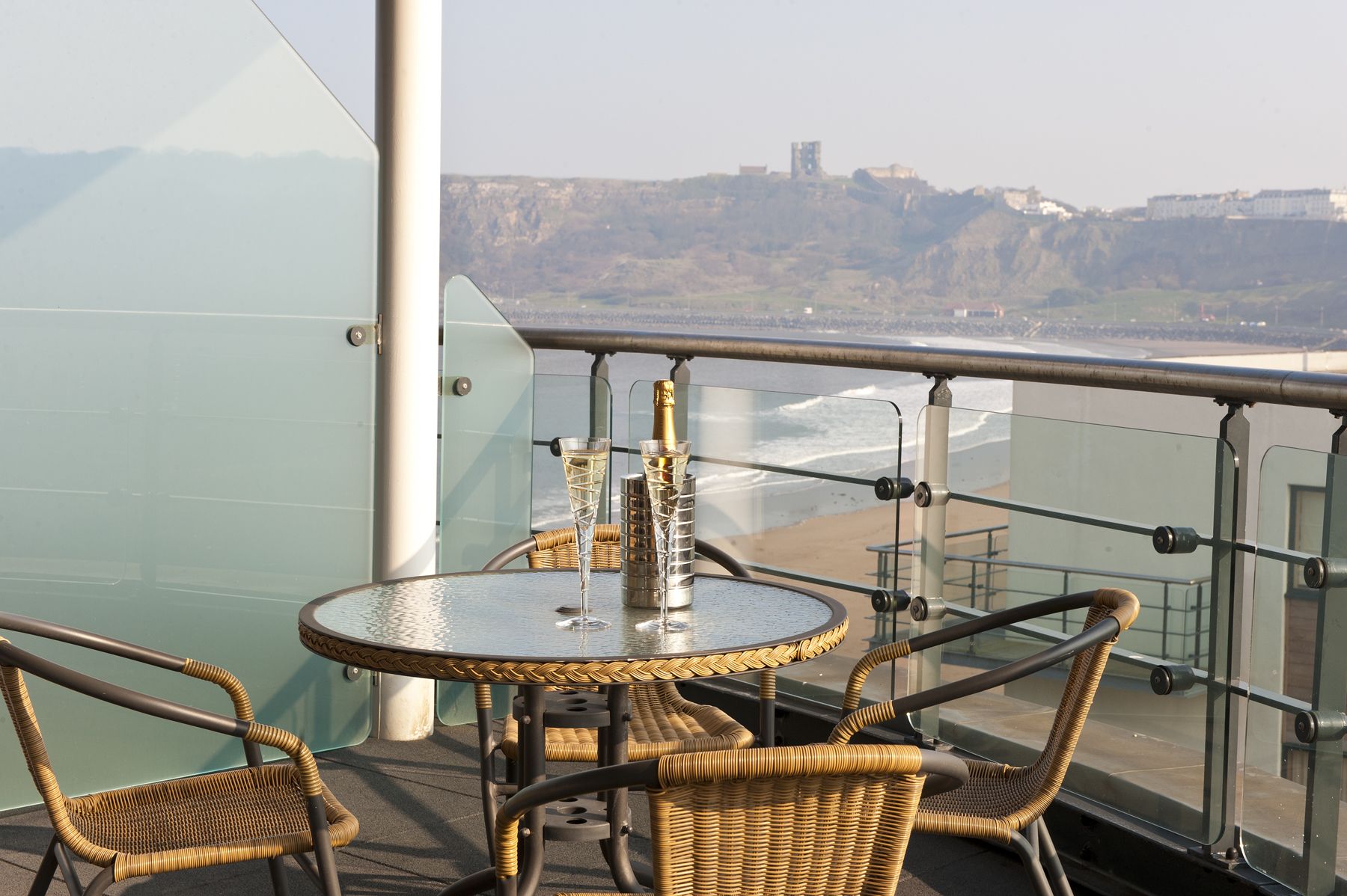 A glass-enclosed balcony with wicker furniture, a round table, and a bottle of champagne.