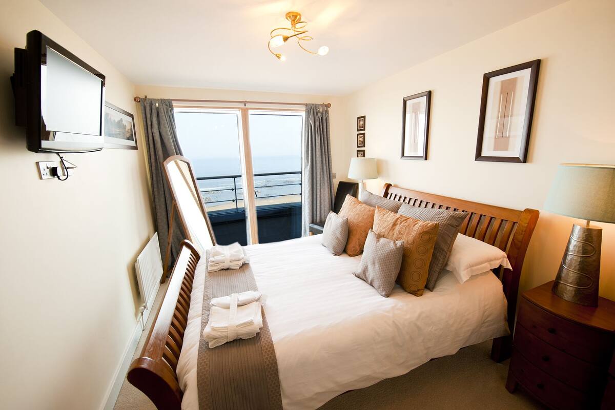 A hotel room with a bed, a TV, a desk, and a view of the sea.