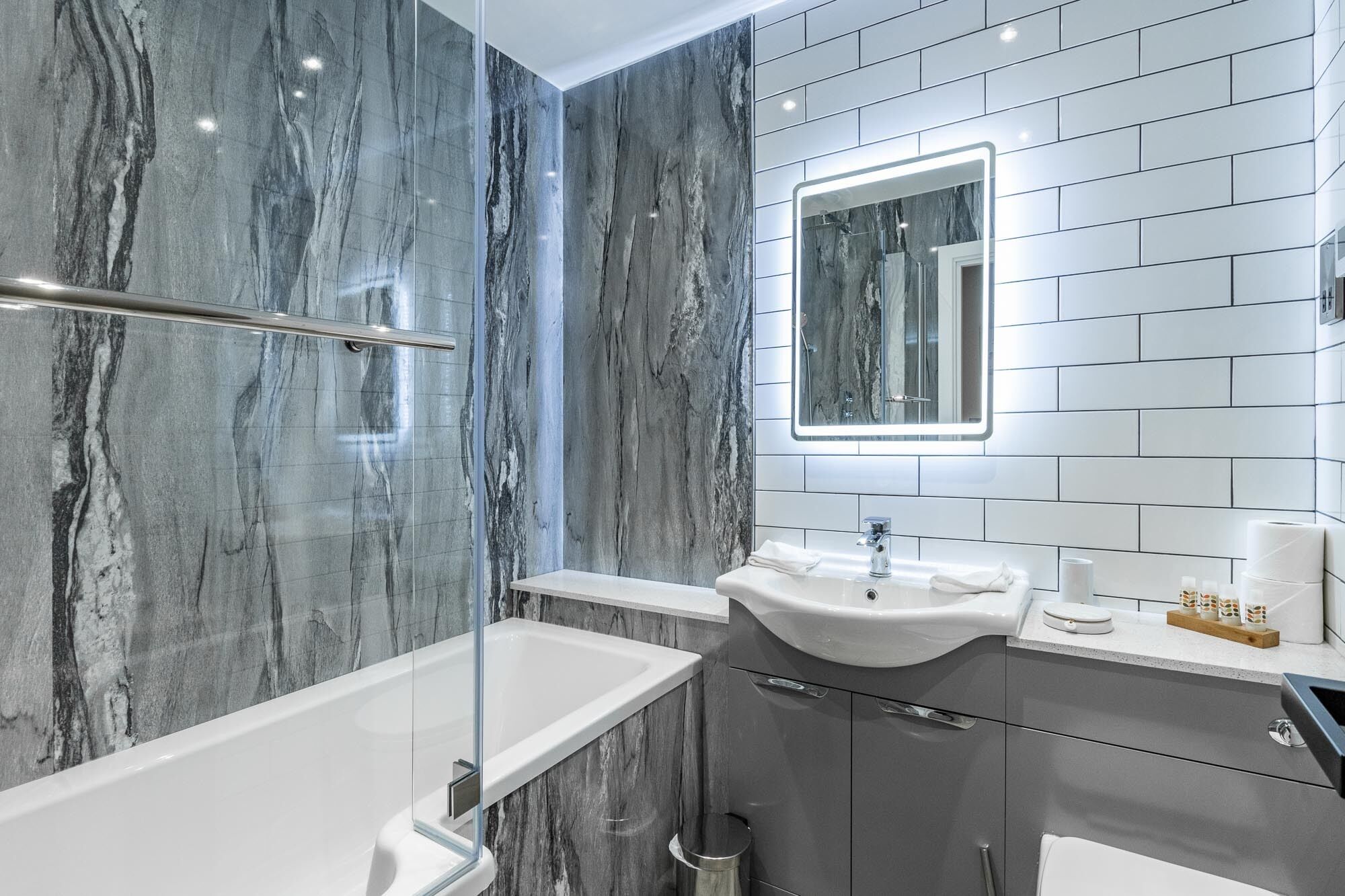 A modern bathroom with a marble wall, a glass-enclosed shower, a white sink, and a mirror with a lighted frame.