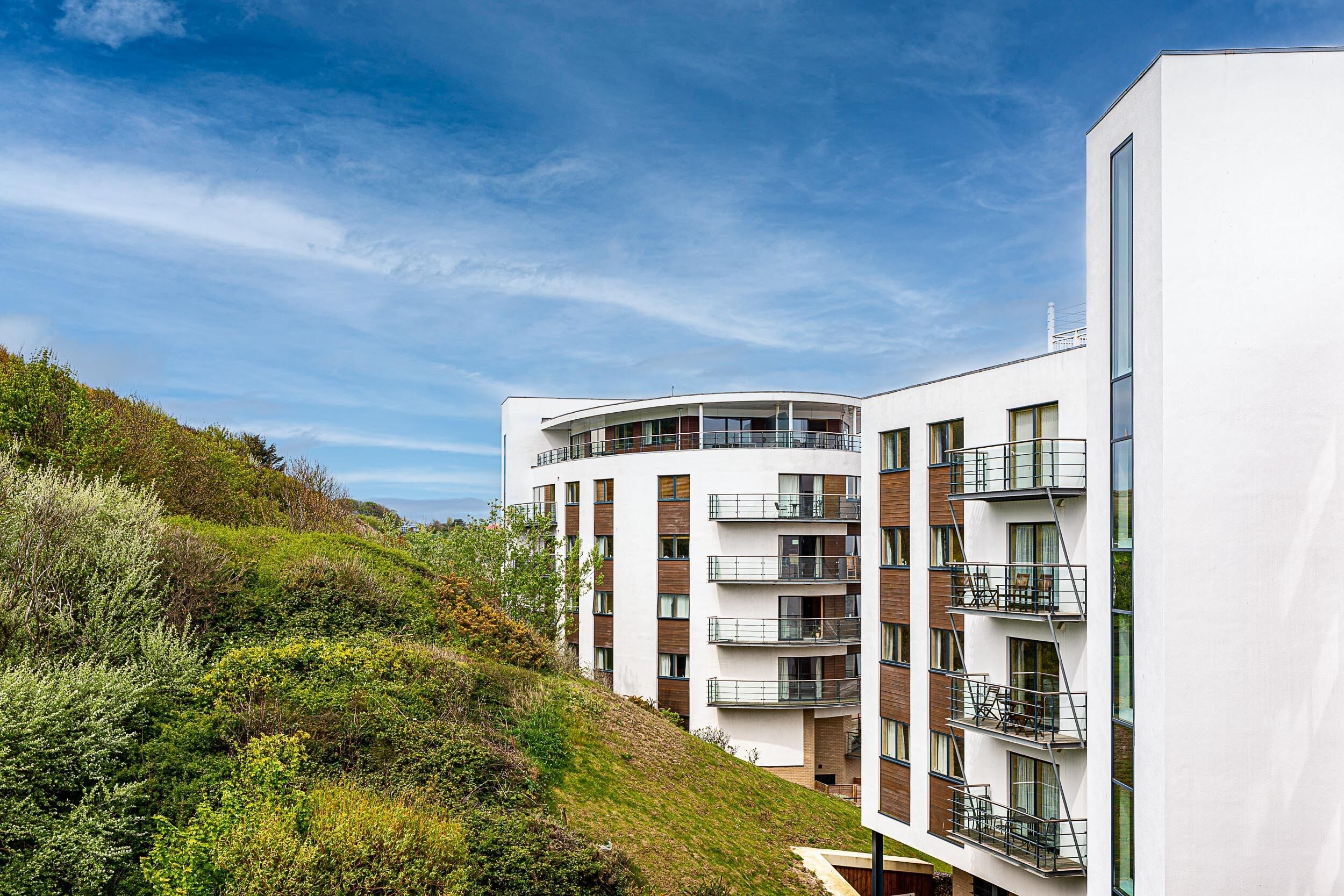 A modern multi-story building with balconies, surrounded by greenery and a clear sky.