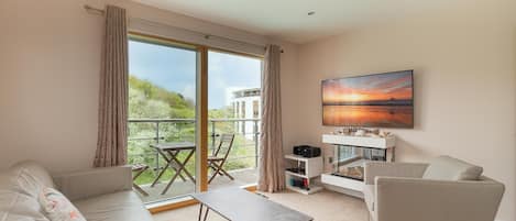 A modern living room with a sofa, a coffee table, and a chair. There is a balcony with a table and chairs, and a painting on the wall.