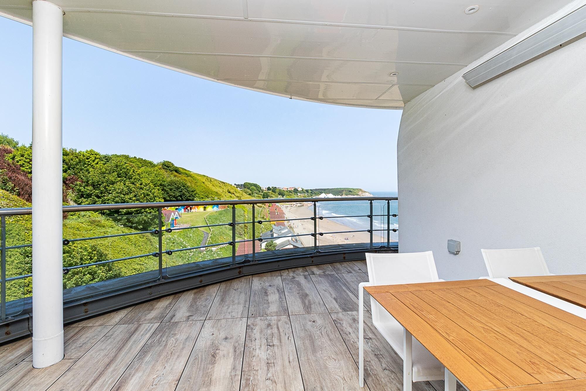 A balcony with a wooden floor, white chairs, and a table. It overlooks a beach and a hilly landscape.
