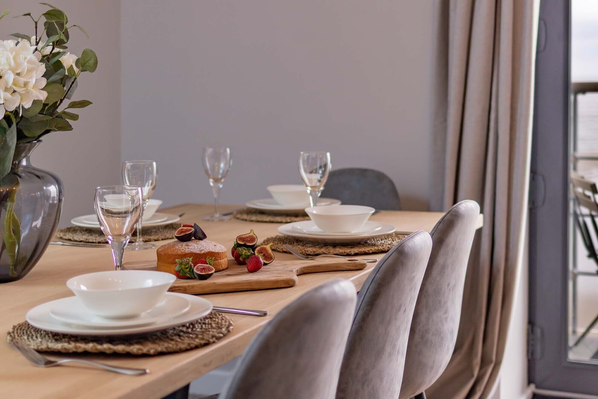A dining table set with white plates, glasses, and a centerpiece of a loaf of bread with strawberries and figs.