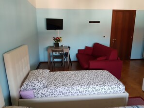 A bedroom with a bed, a desk with a chair, a red sofa, a television on the wall, and a wooden door. - New Life (Mestre)