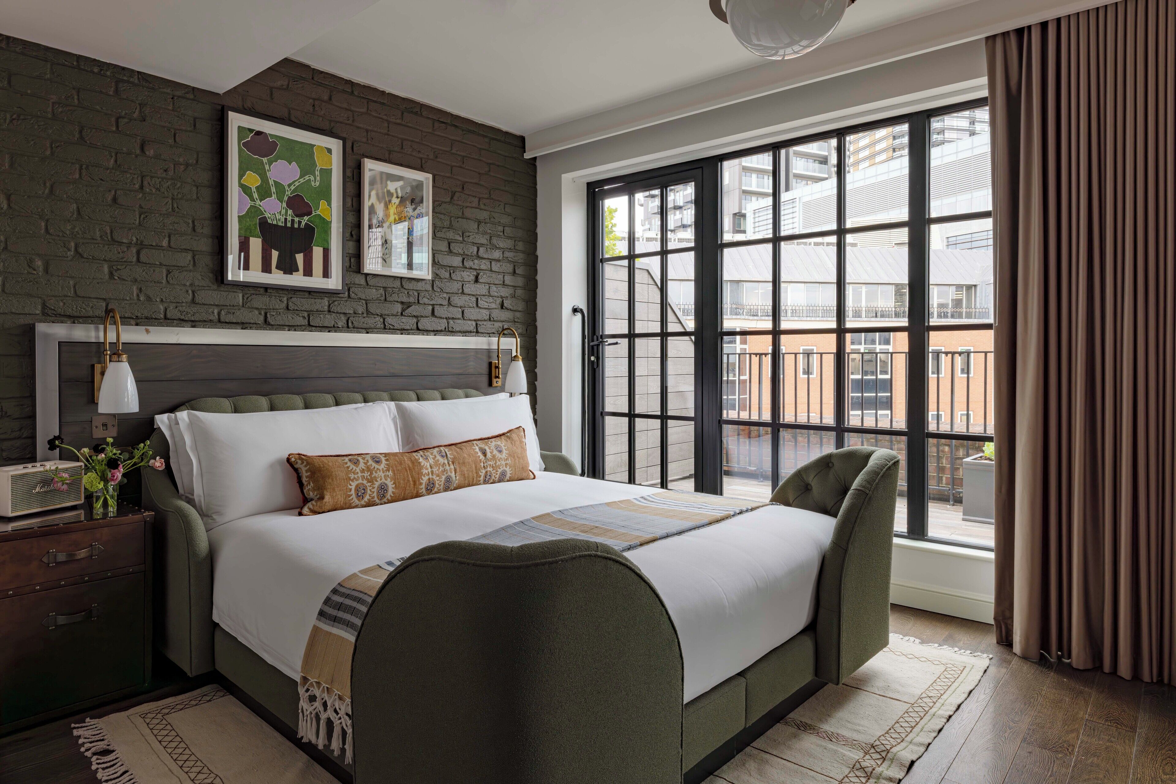 A modern bedroom with a large bed, a dresser, a nightstand, a window with a view of the city, and a brick accent wall.