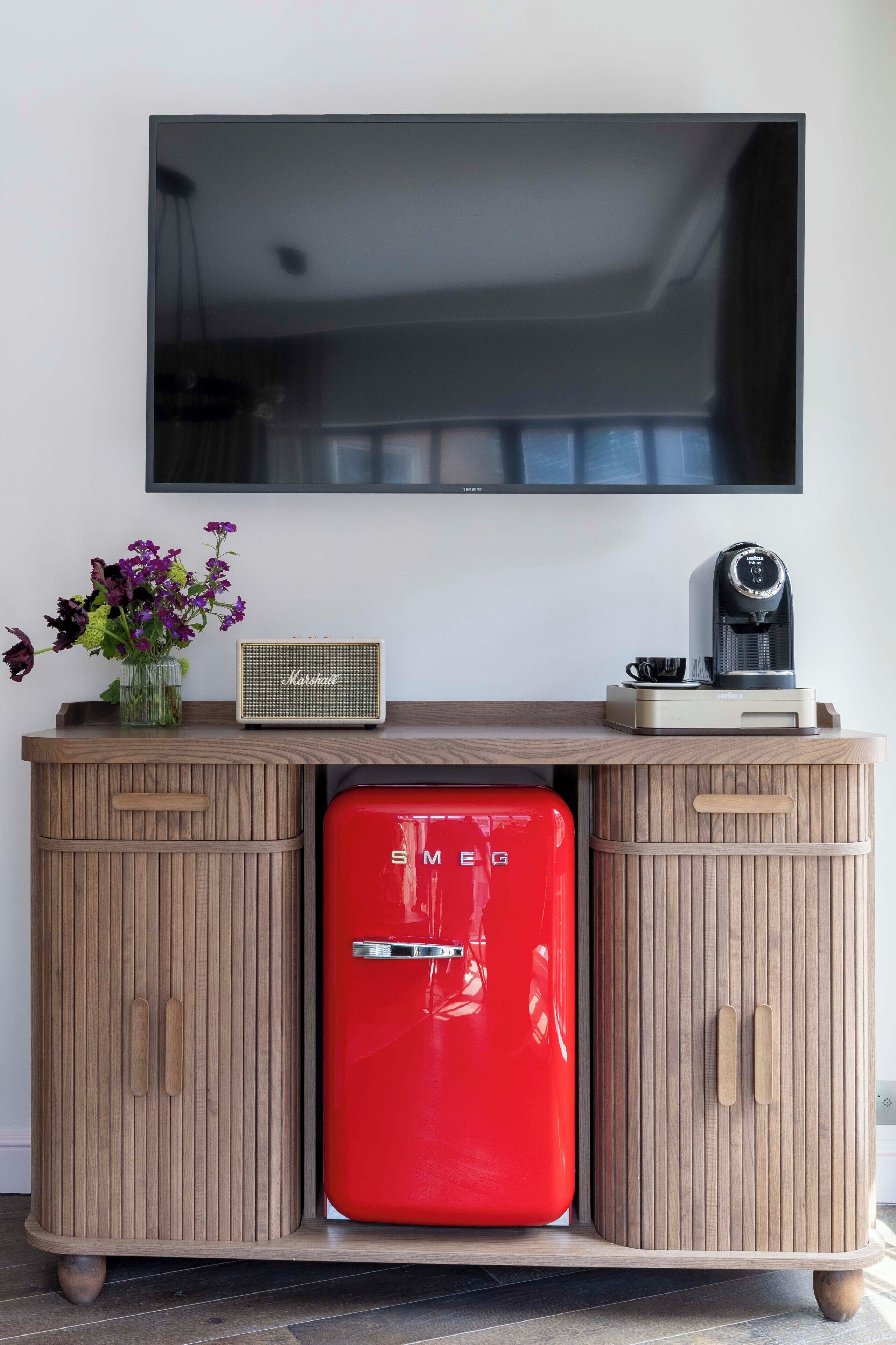 Una sala de estar moderna con un refrigerador rojo, una cafetera y un televisor de pantalla plana montado en la pared.