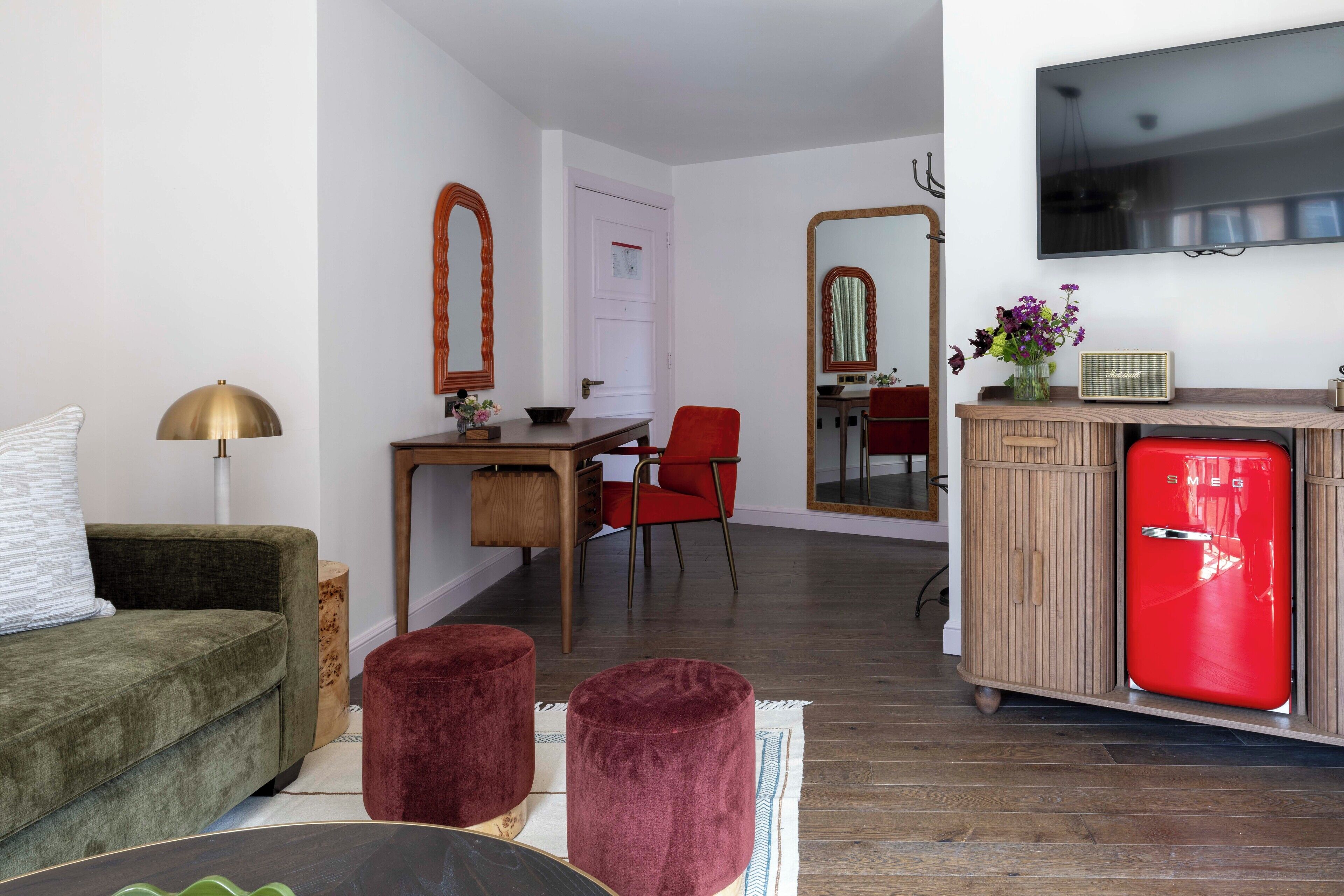 A modern living room with a green sofa, a red chair, a wooden desk, a red refrigerator, and a TV mounted on the wall.