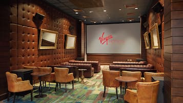 Een gezellige zithoek met lederen zitplaatsen, kleine tafels en een groot scherm waarop het Virgin Hotels logo staat.