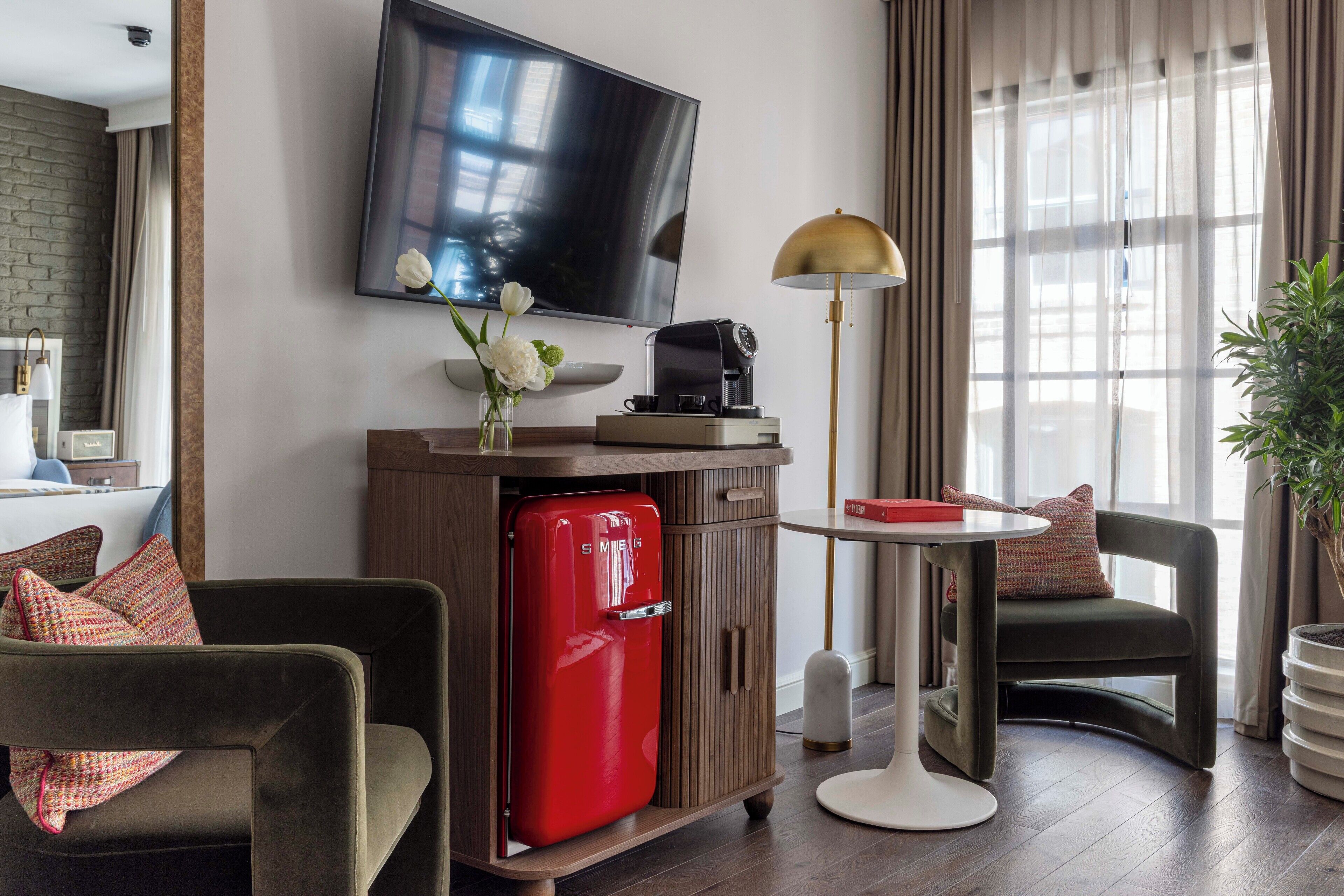 A modern hotel room with a flat-screen TV, a red refrigerator, a coffee maker, a small table, and a comfortable seating area.
