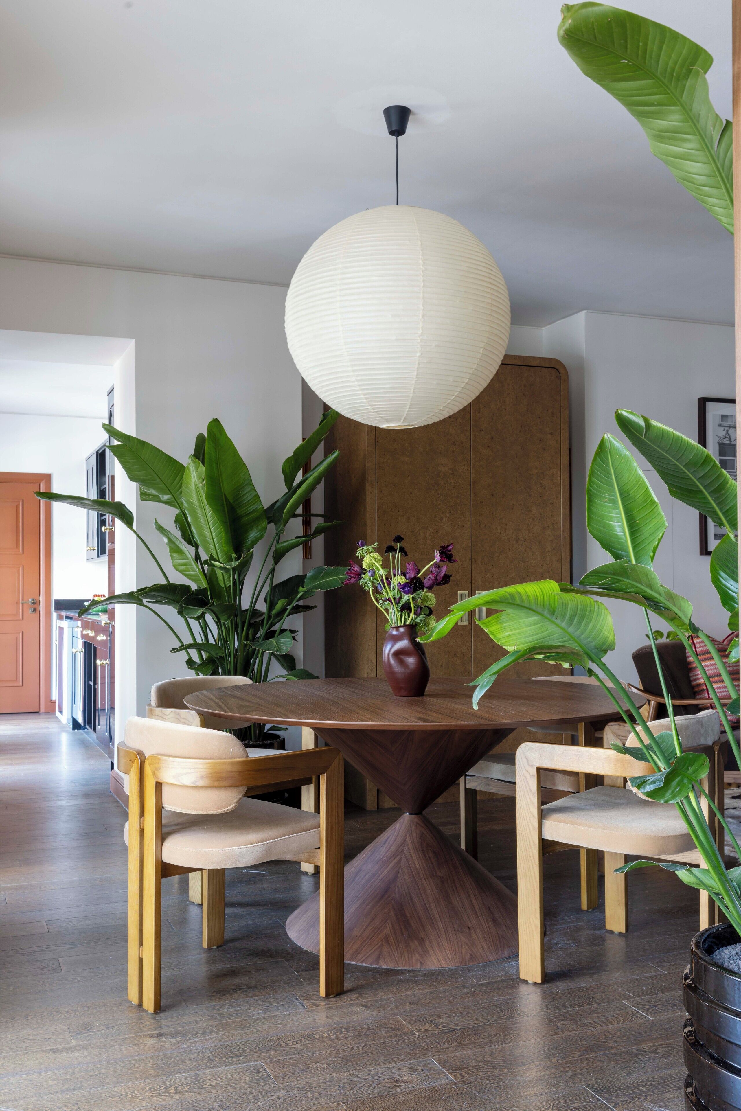 Un coin repas avec une table ronde en bois, quatre chaises, un vase de fleurs et de grandes plantes vertes.