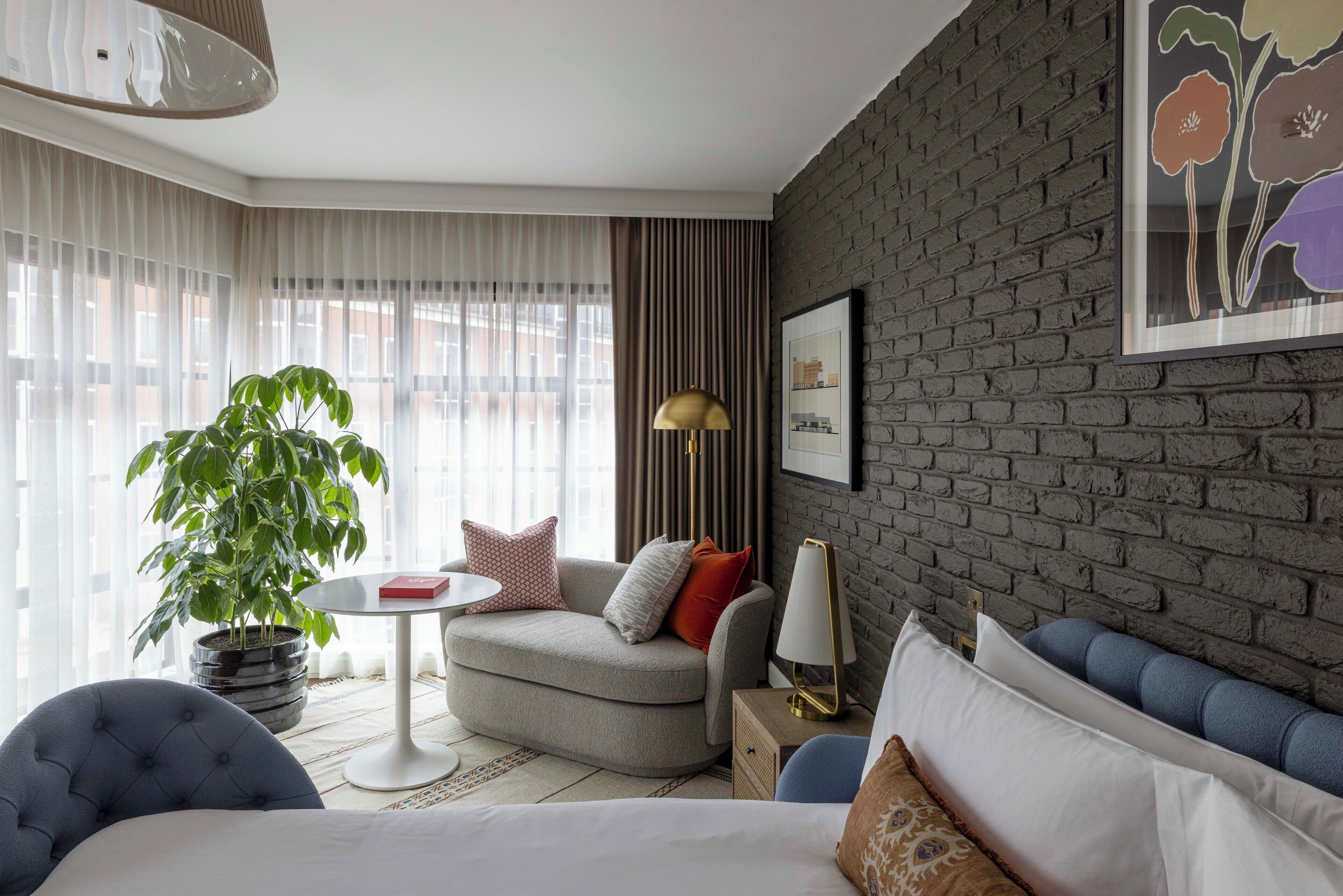 A modern hotel room with a grey brick wall, a large window with sheer curtains, a grey sofa, a round coffee table, a potted plant, and a bed with white bedding.