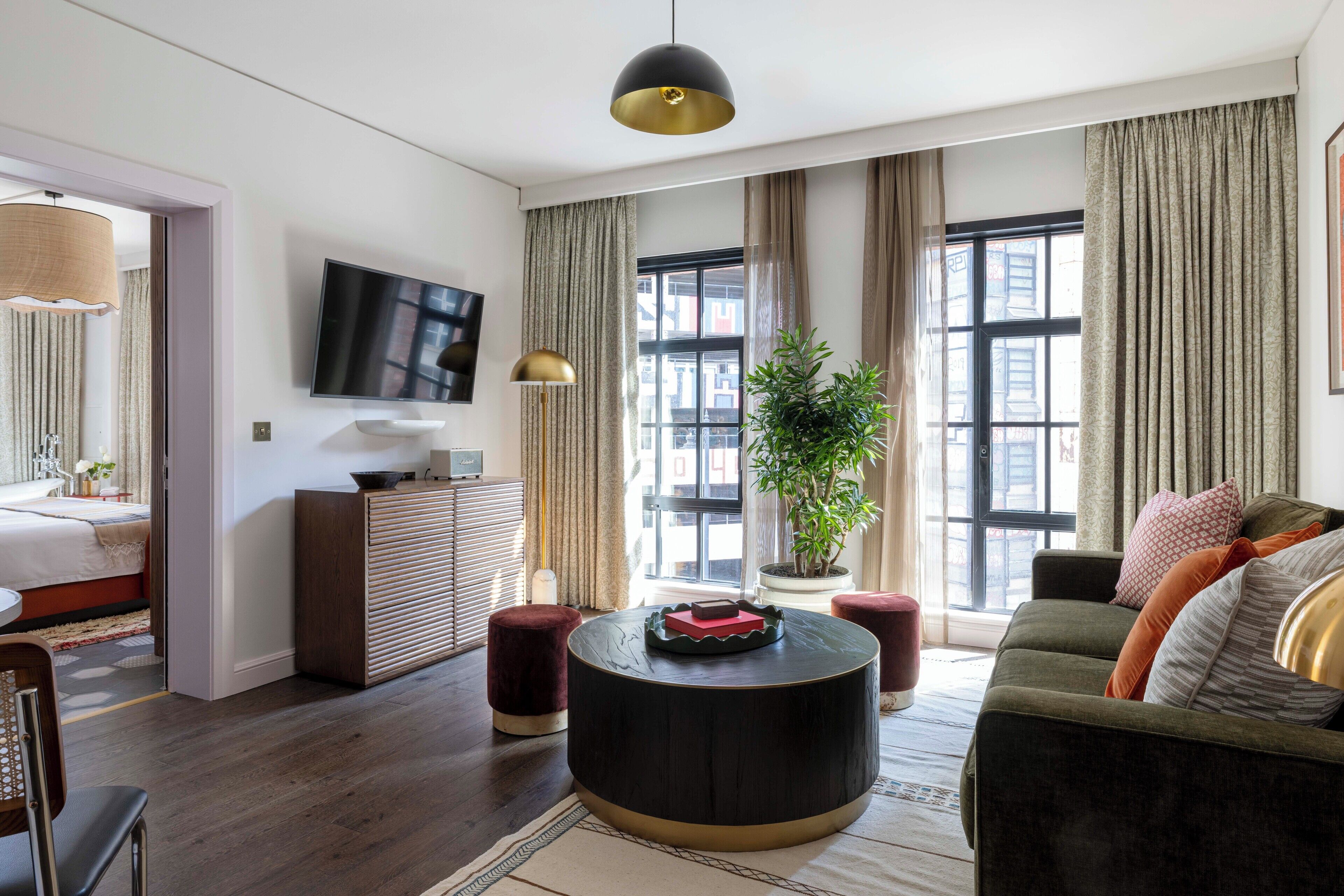 A modern living room with a sofa, a round coffee table, a TV mounted on the wall, and a large window with curtains.
