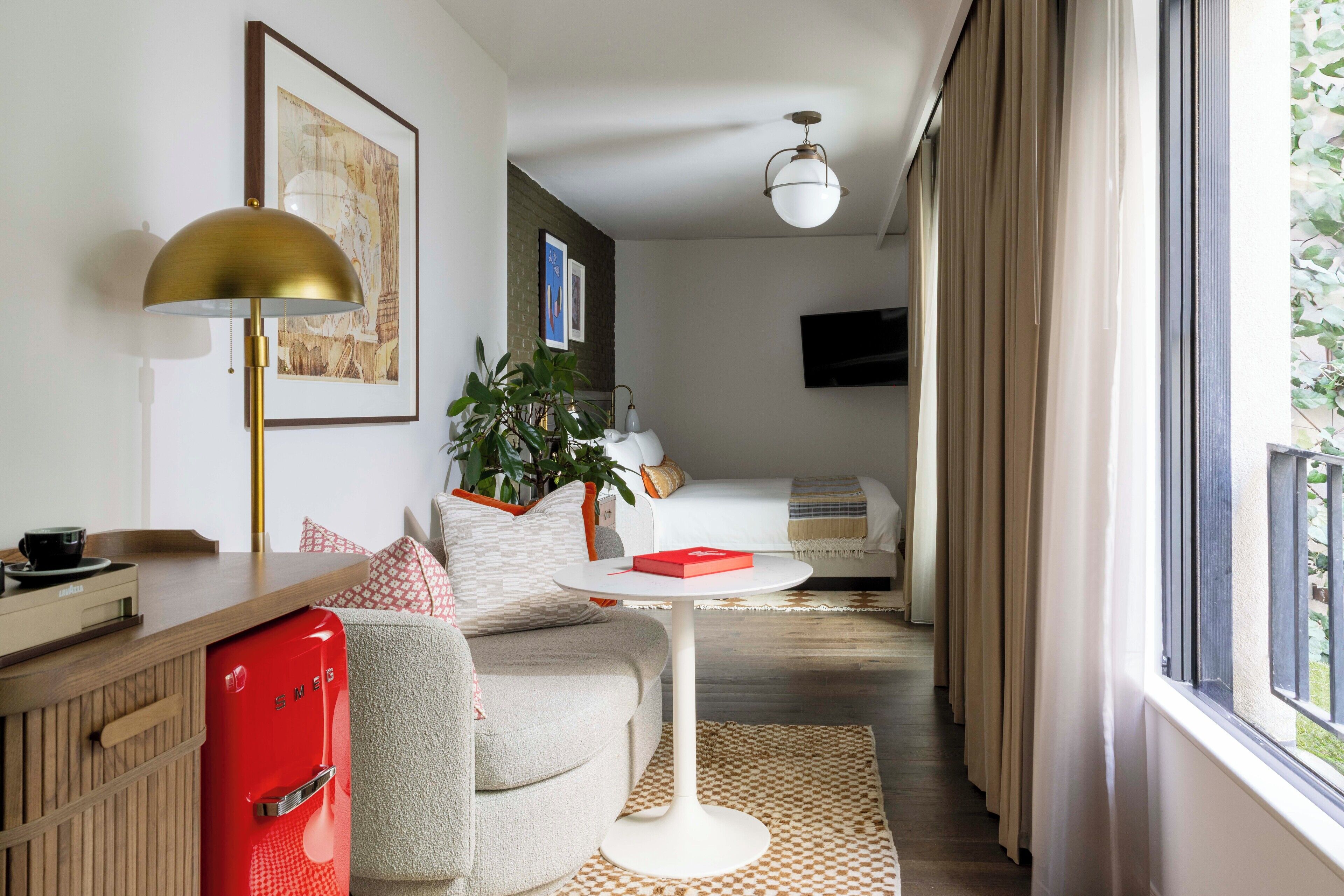A modern hotel room with a sofa, a small round table, a red minibar, a lamp, a plant, and a TV mounted on the wall.