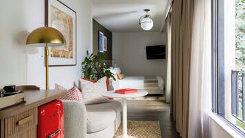 A modern hotel room with a sofa, a small round table, a red minibar, a lamp, a plant, and a TV mounted on the wall.