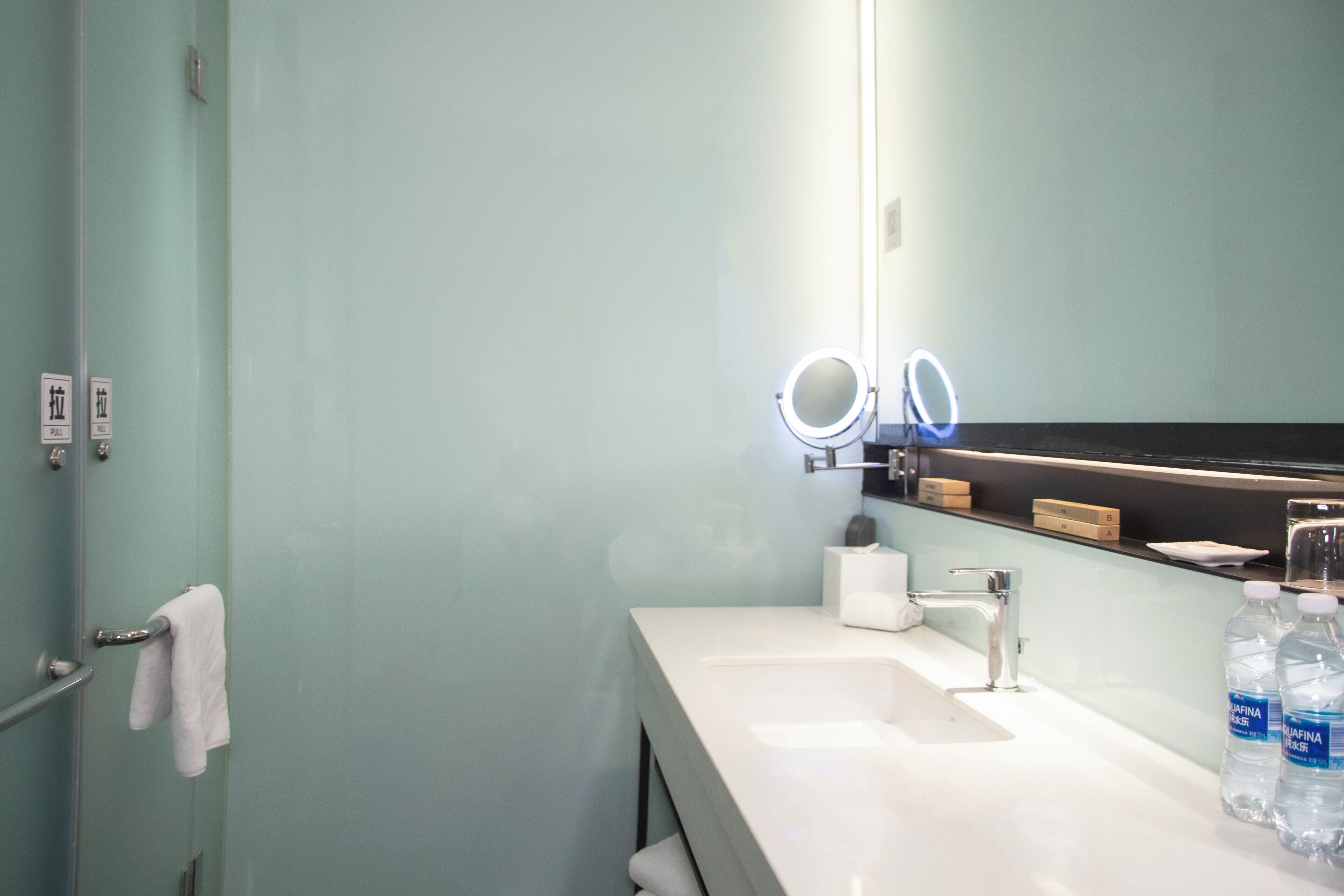 Une salle de bain avec un lavabo blanc, un miroir doté d’anneaux lumineux, un porte-serviettes et deux bouteilles d’eau.