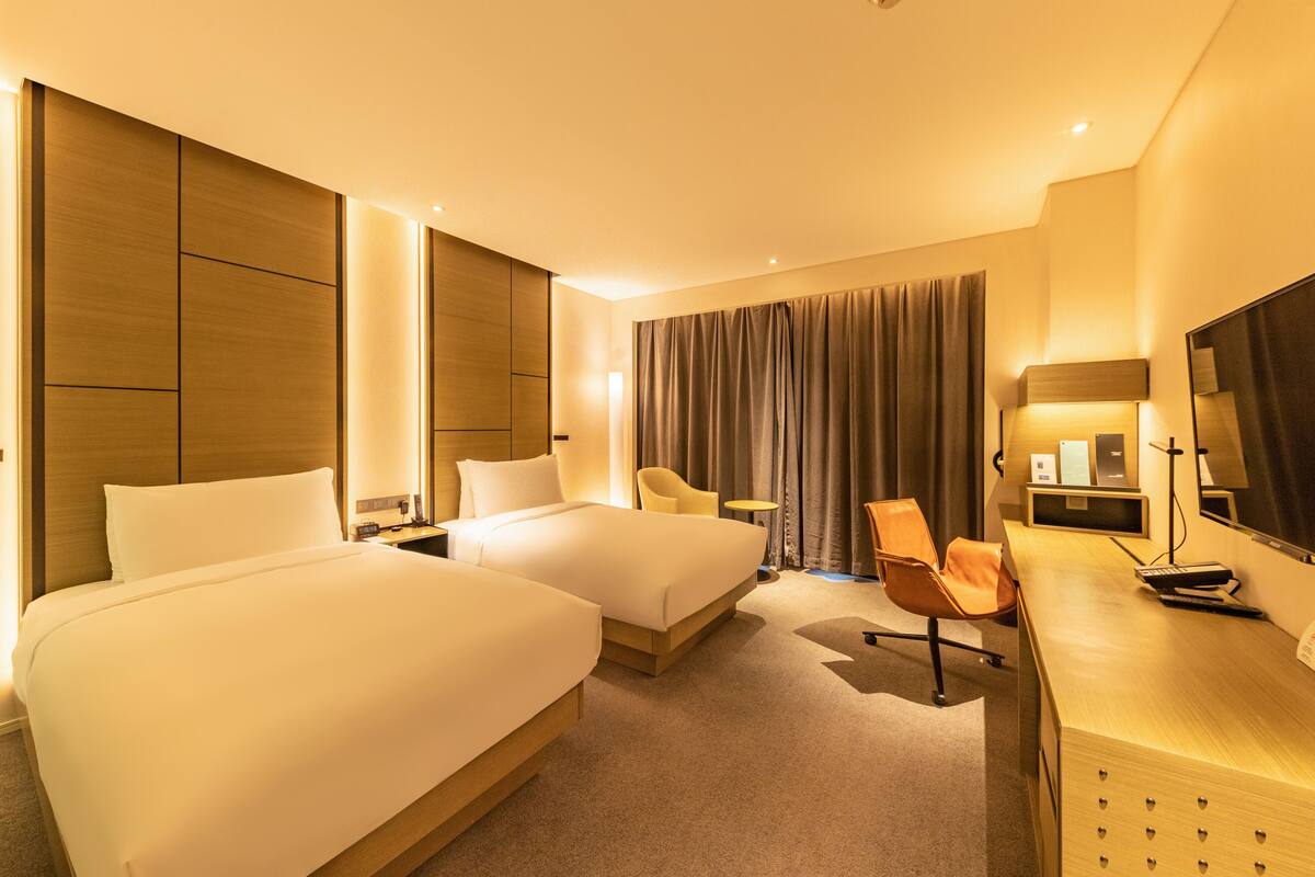 A hotel room with two beds, a desk, a chair, a TV, and a large window with curtains.