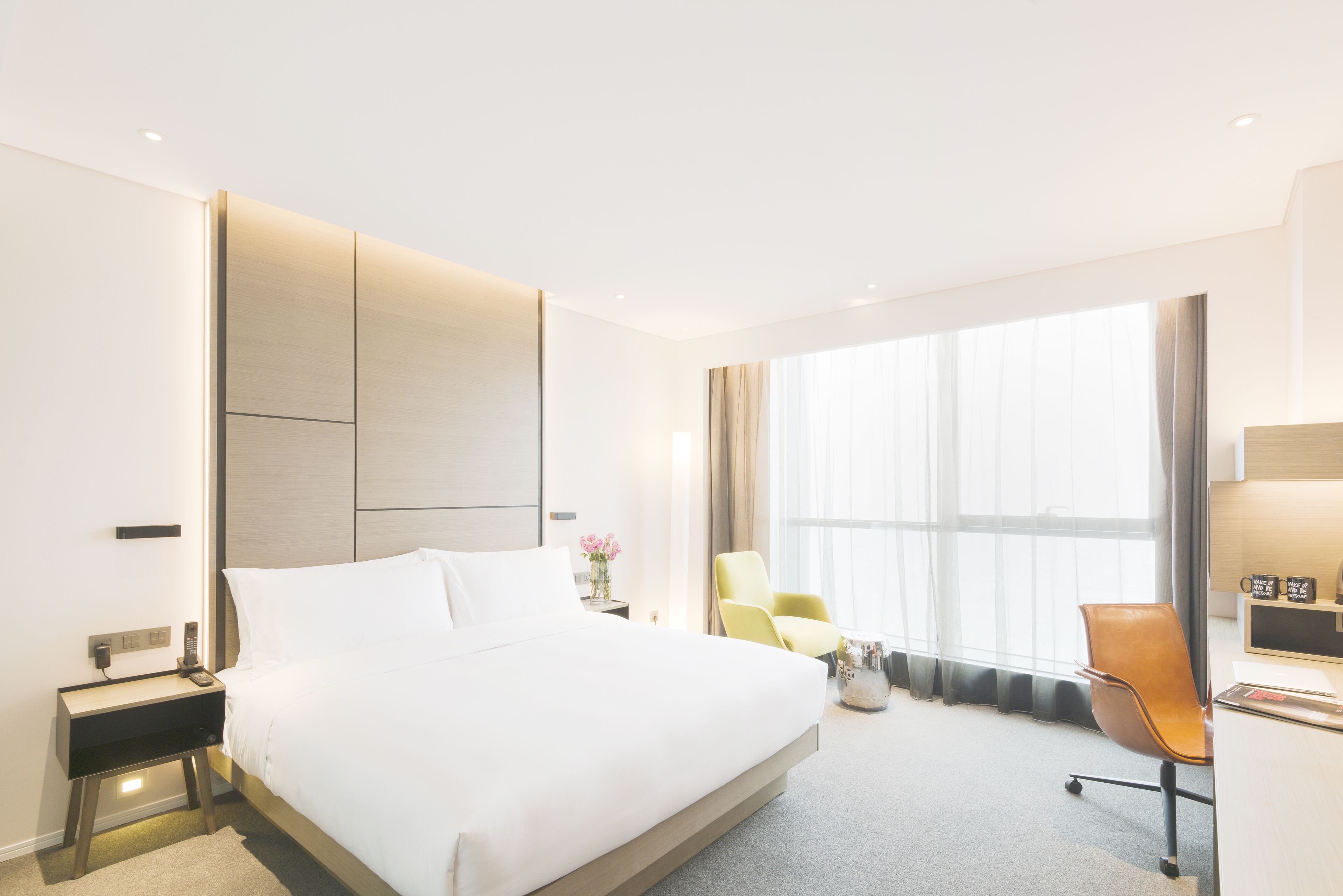 A modern hotel room with a large bed, a desk, a chair, and a large window.