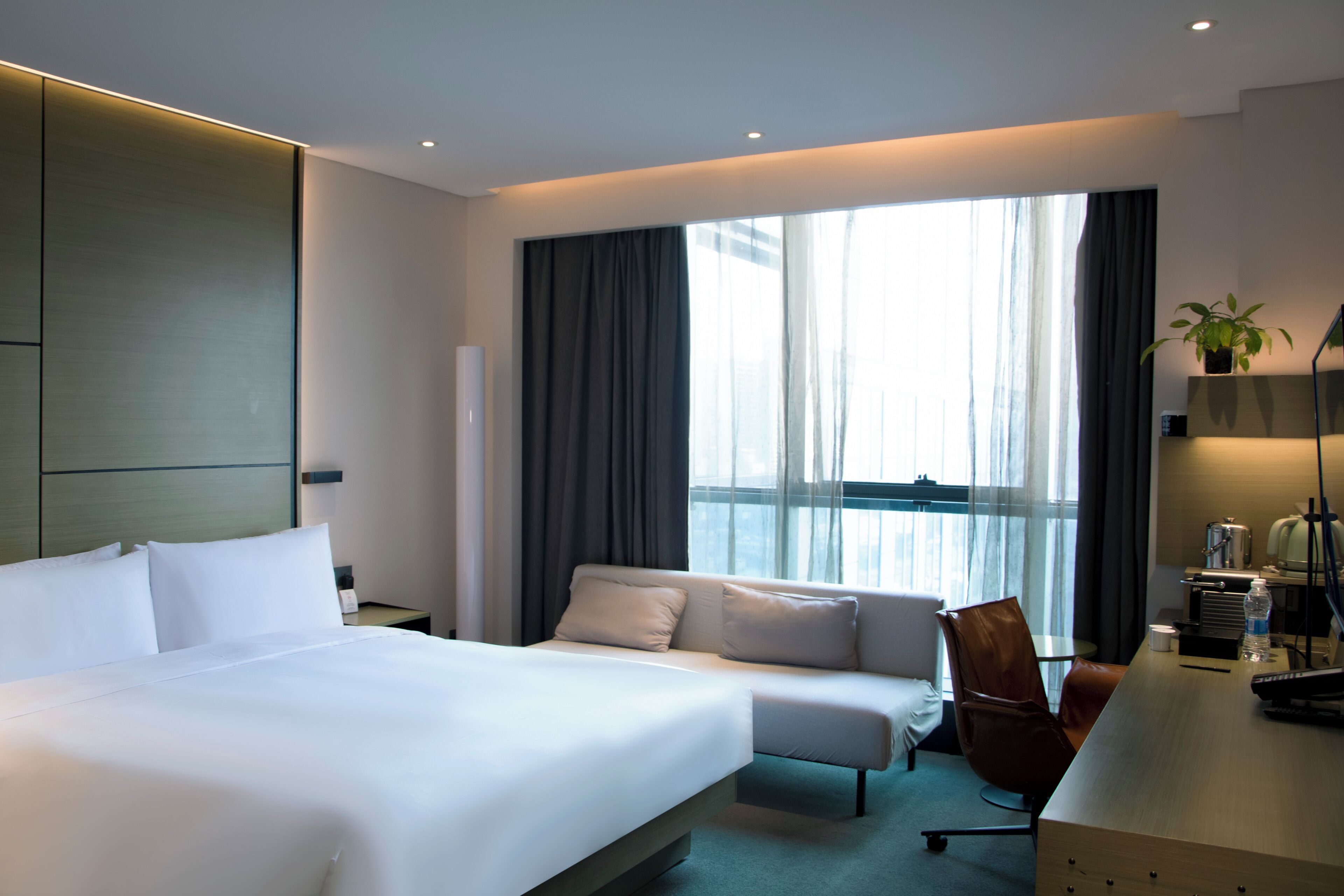 A hotel room with a bed, a sofa, a desk, and a large window with curtains.