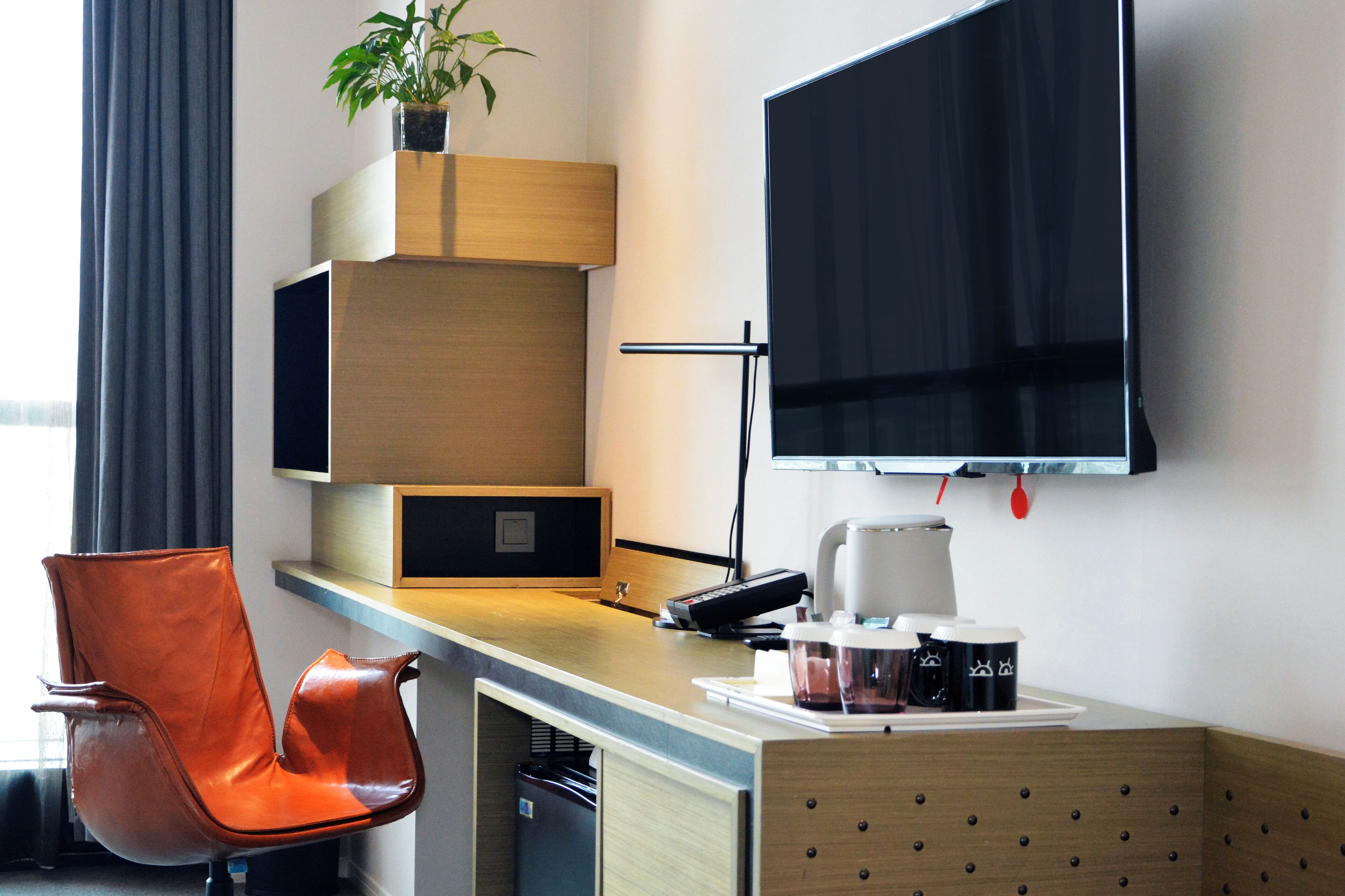 A modern hotel room with a wooden desk, a leather chair, a flat-screen TV, a coffee maker, and a potted plant.