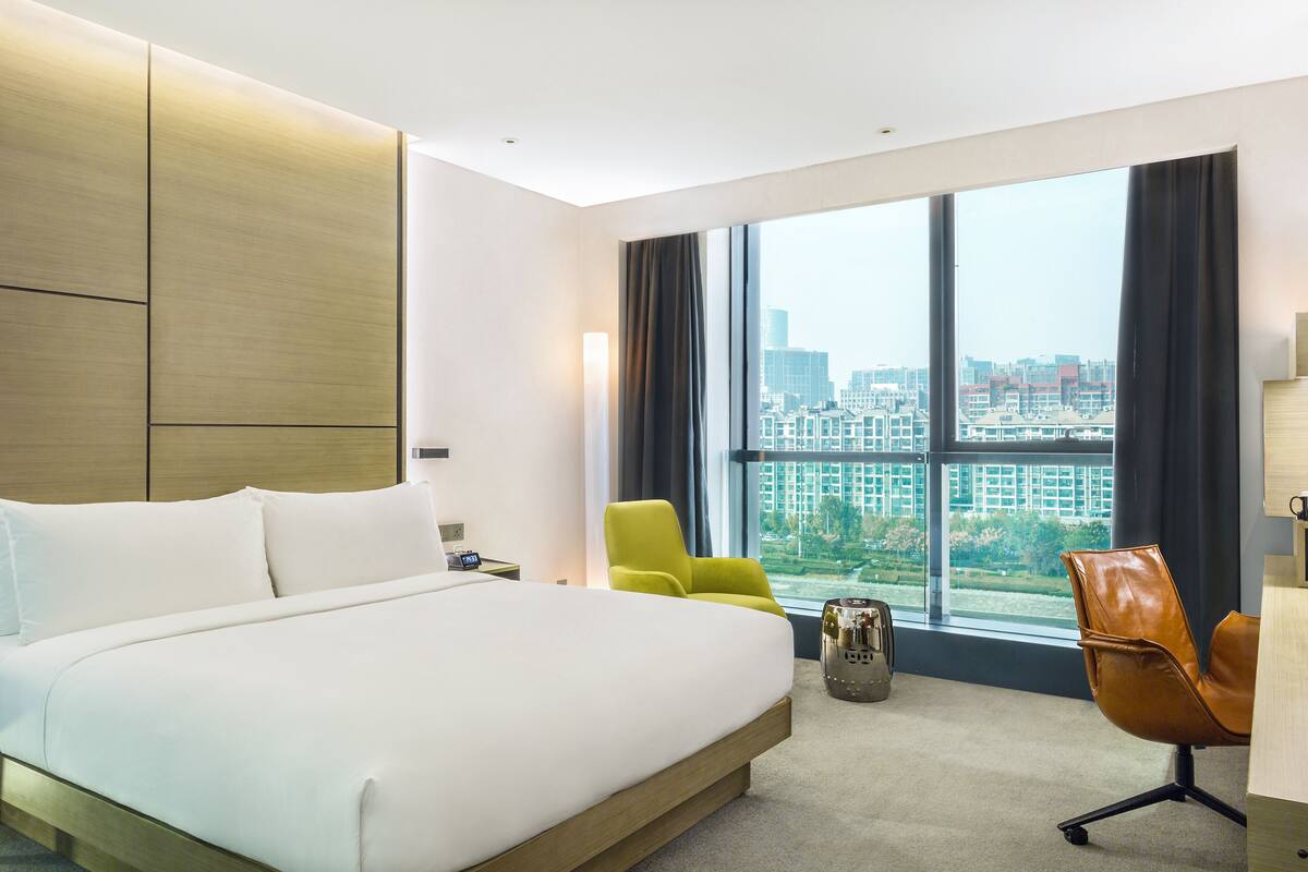 A modern hotel room with a large bed, a desk, a chair, and a view of the city through large windows.