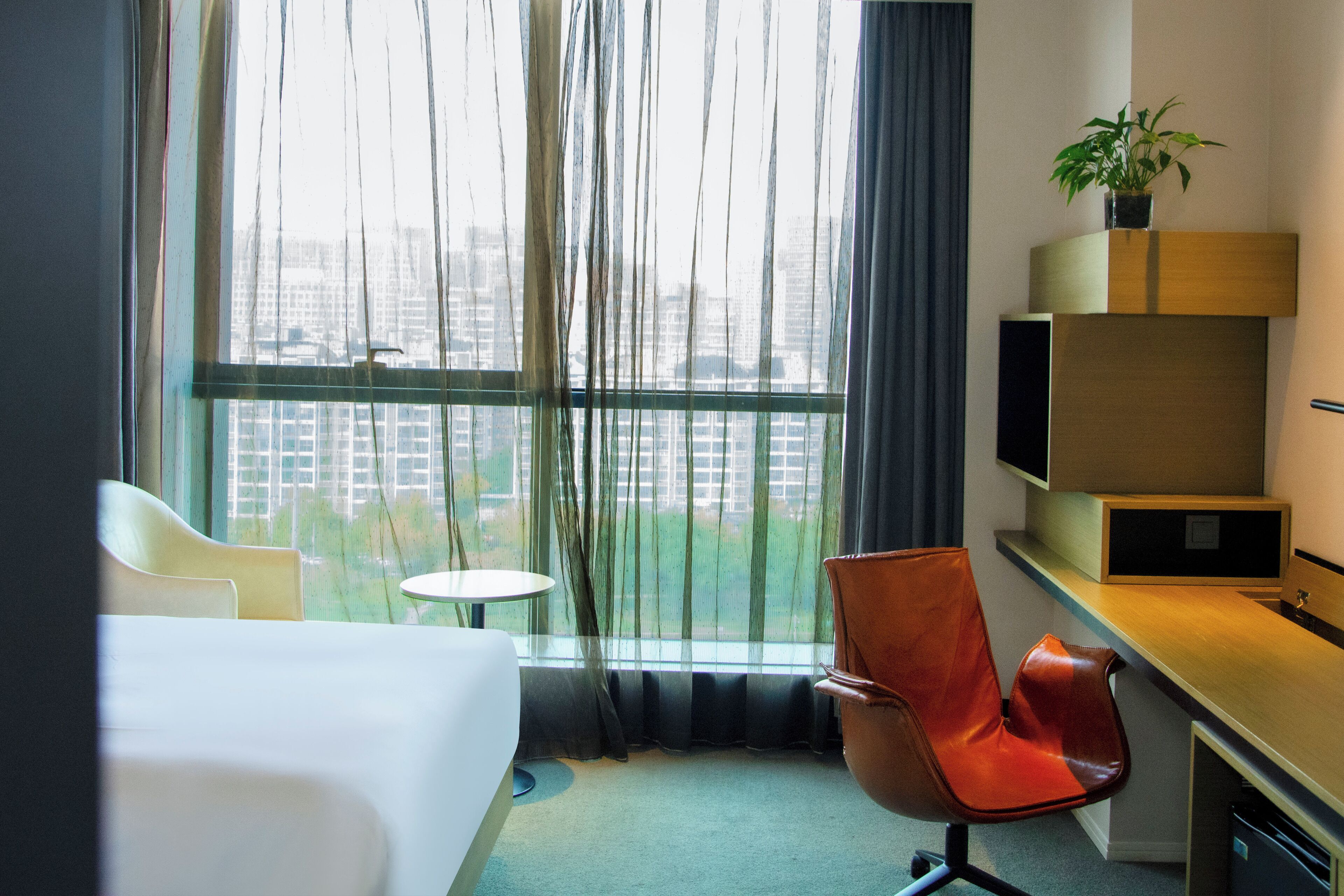 A hotel room with a bed, a desk, a chair, a small table, and a large window with curtains.