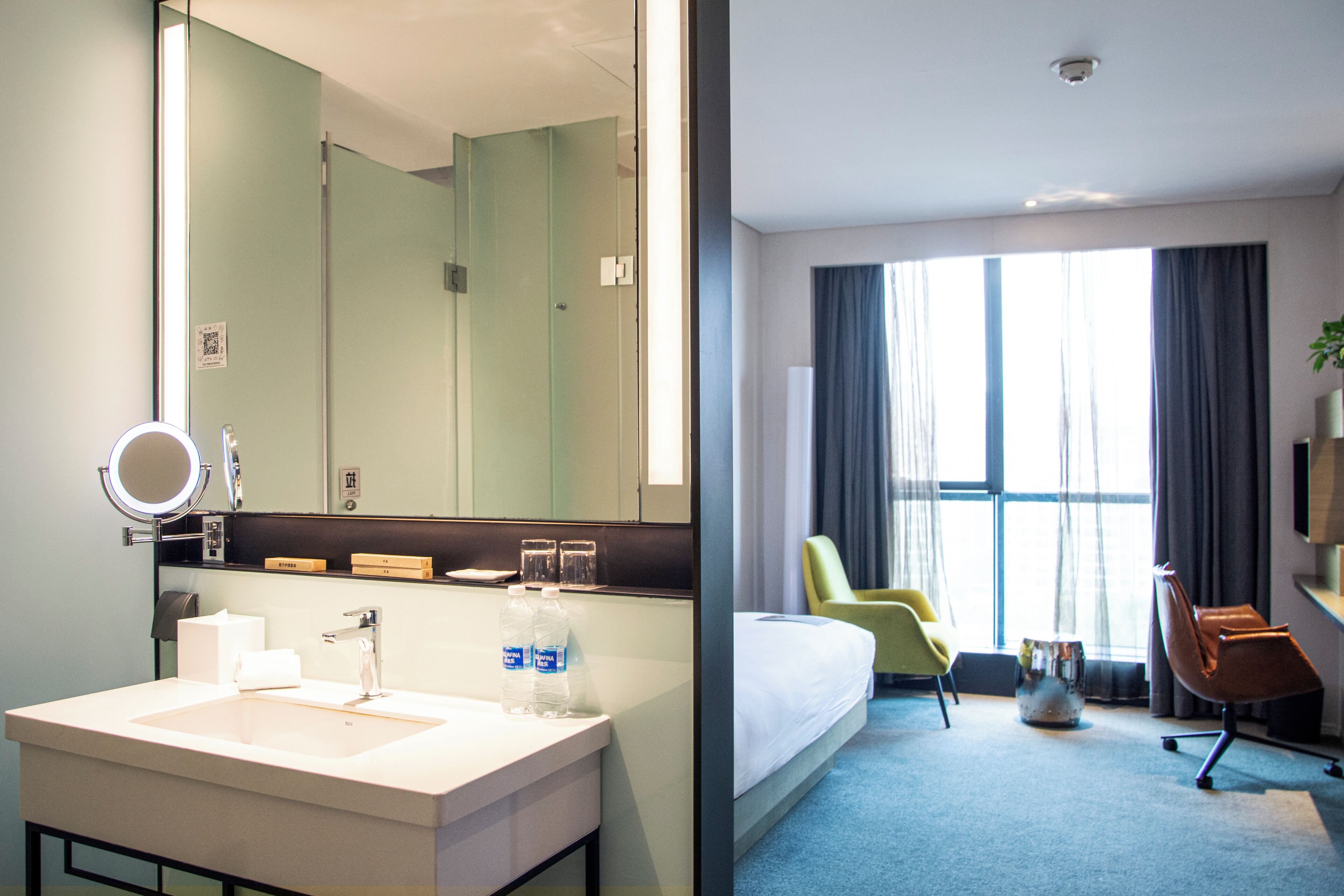 Hotelkamer met een grote spiegel, een wastafel, een bed, een stoel en een raam met gordijnen.