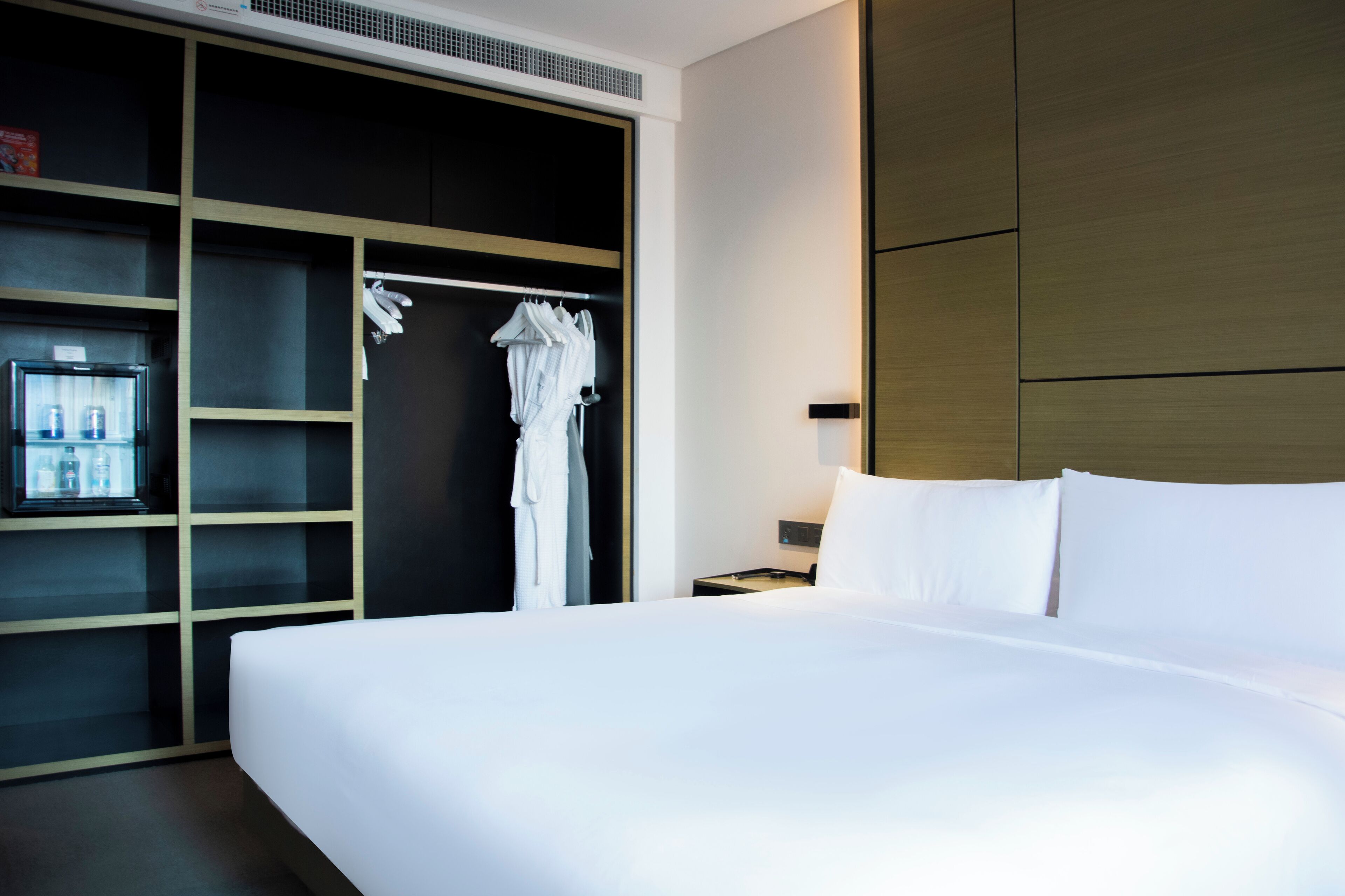 A hotel room with a large bed, a wardrobe with shelves, and a small table with a lamp.
