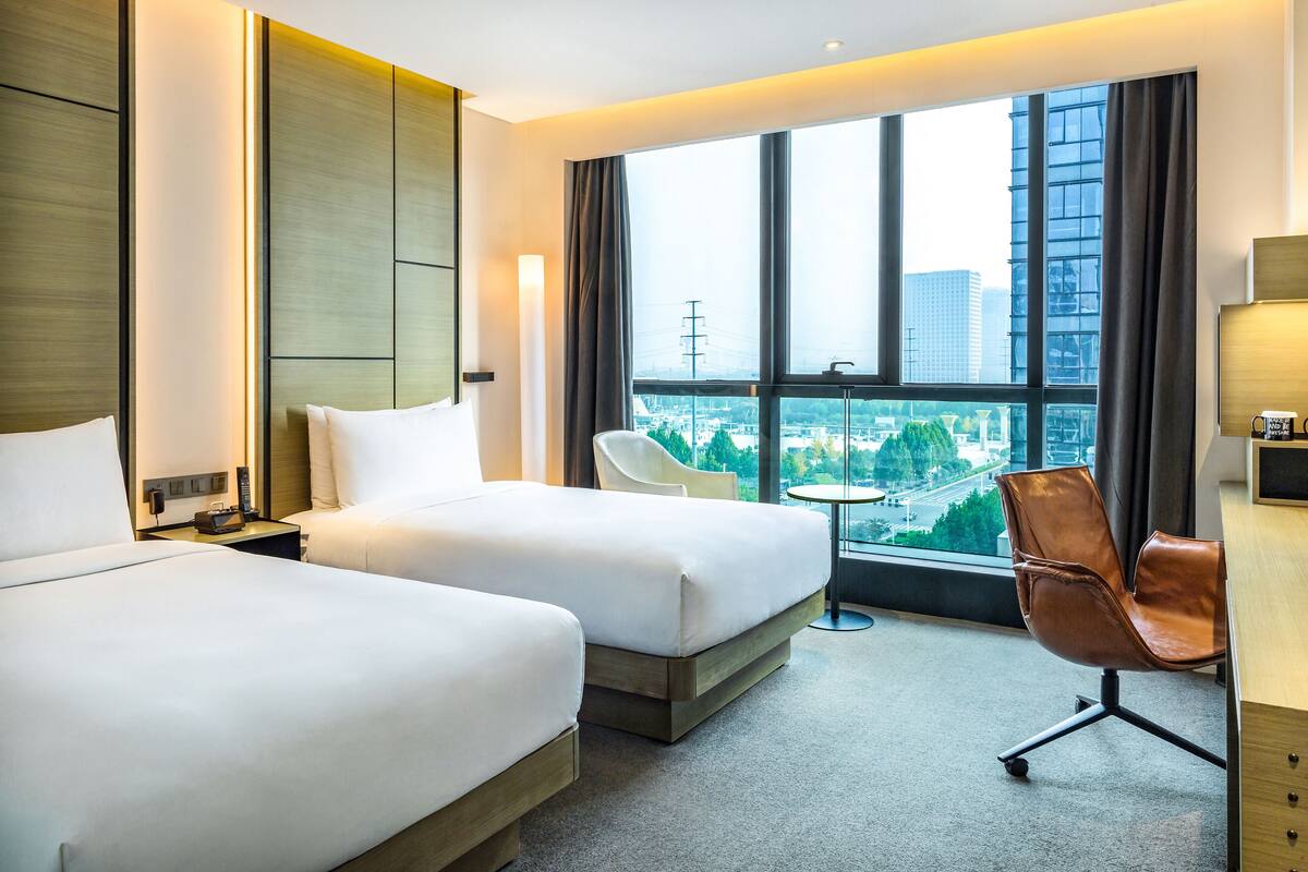 A hotel room with two beds, a desk, a chair, and a large window offering a city view.